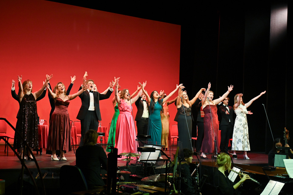 A group of singers performing on stage, with an orchestra below them, with a red background.
